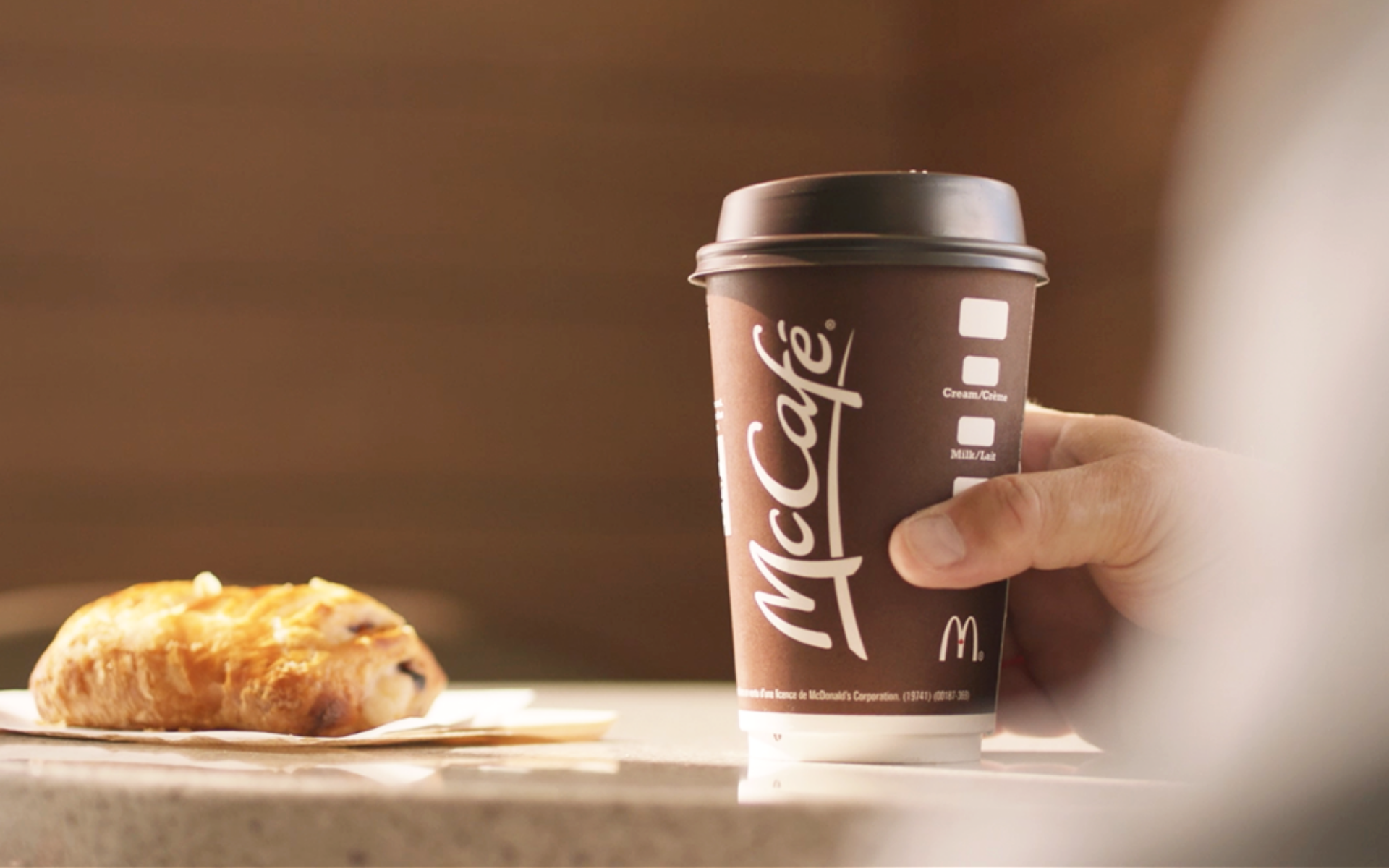 Hand holding McCafe premium roast specialty coffee. A chocolate pastry is next to the coffee cup on the table.